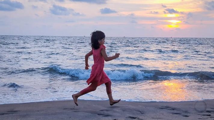 A young girl in a red dress running along the shoreline at sunset, the sun low on the water behind her.