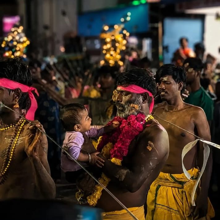A festival at night in Villivakkam: a man carrying a baby who is looking up at him, lights blurred behind them.